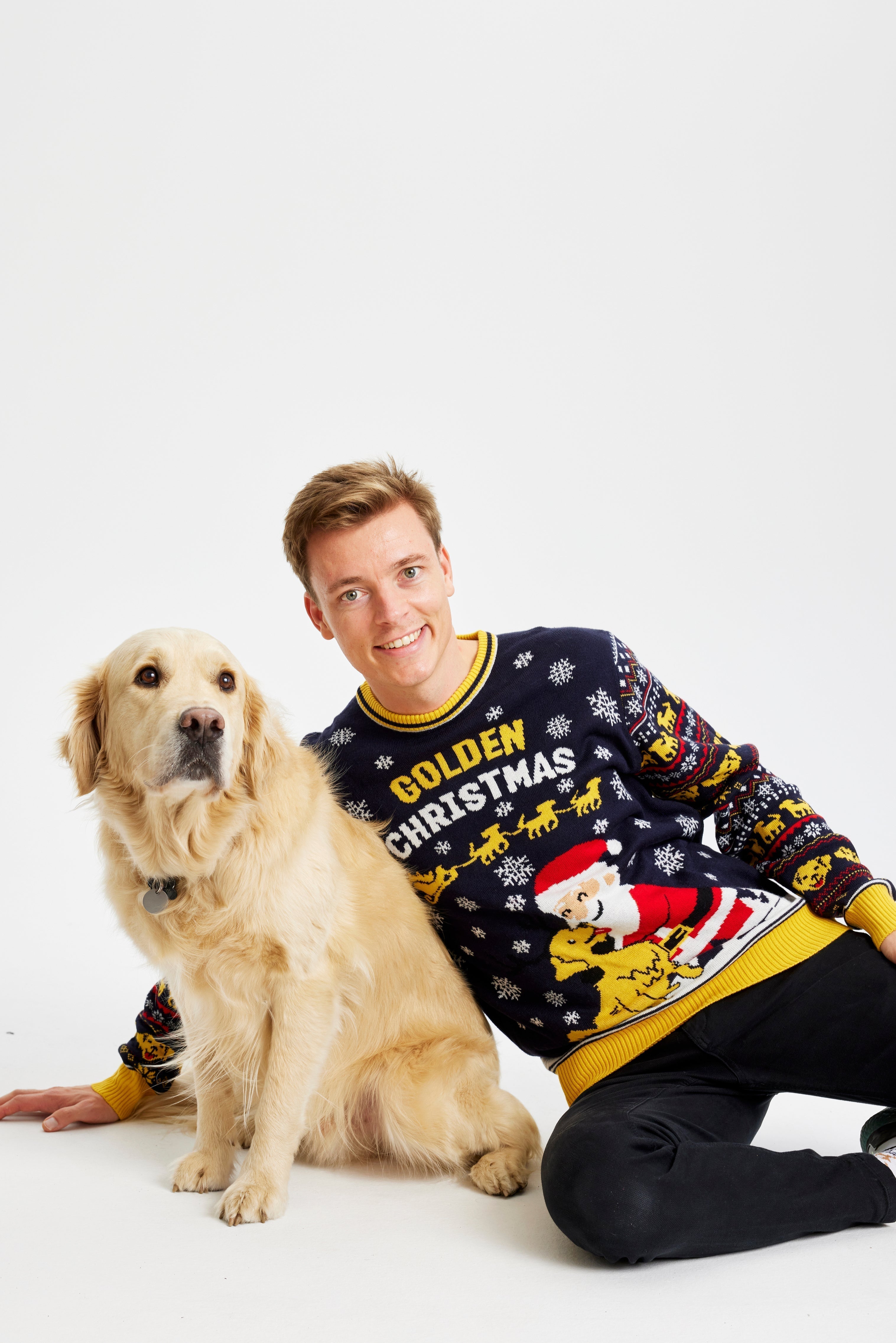 Un homme souriant se couche avec un chien vêtu d'un pull de Noël jaune et noir portant l'inscription